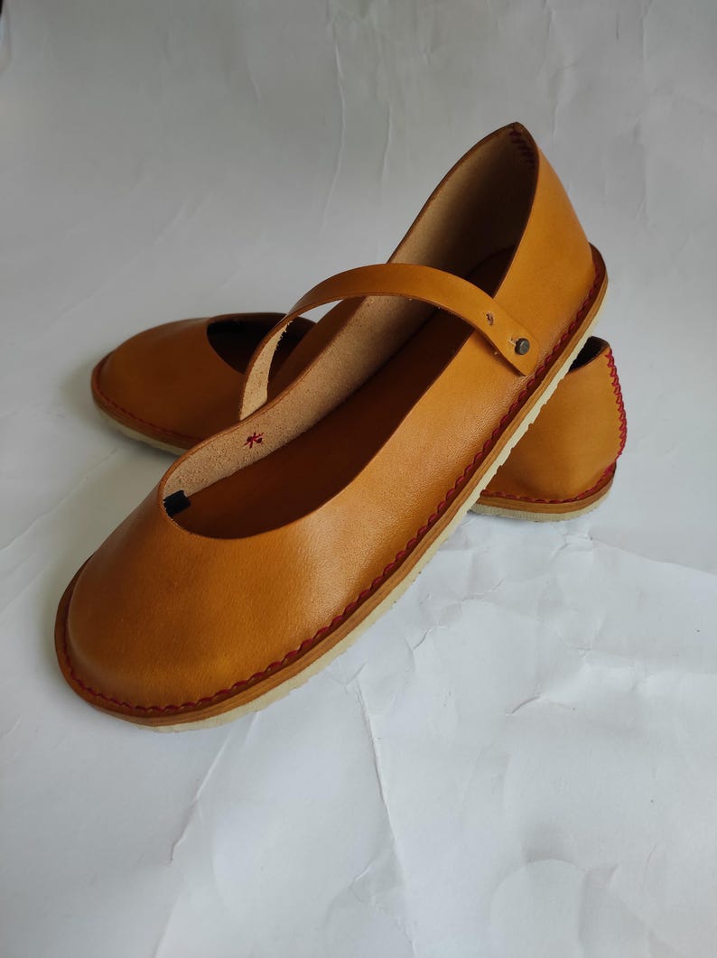 Pode incluir: Um par de sapatos baixos de couro castanho com um detalhe de costura vermelha em volta da sola. Os sapatos t&ecirc;m um design simples com uma &uacute;nica tira na parte superior.