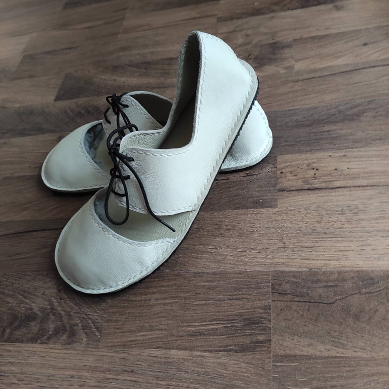 Puede incluir: Un par de zapatos de cuero blanco con un dise&ntilde;o de cordones y un detalle de corte en la parte superior. Los zapatos tienen un detalle de costura negra alrededor de los bordes.