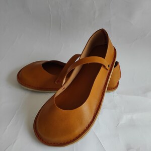 Pode incluir: Um par de sapatos baixos de couro castanho com uma tira na parte superior. Os sapatos t&ecirc;m um detalhe de costura vermelha em volta da borda.