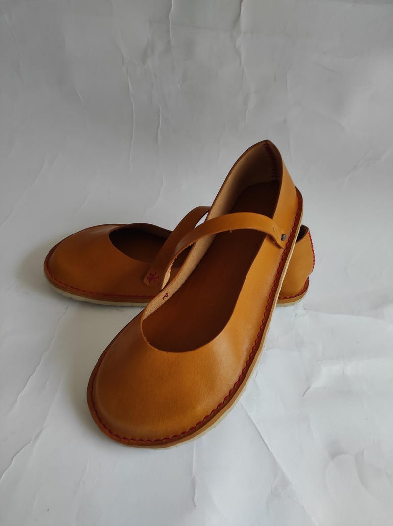 Pode incluir: Um par de sapatos baixos de couro castanho com uma tira na parte superior. Os sapatos t&ecirc;m um detalhe de costura vermelha em volta da borda.