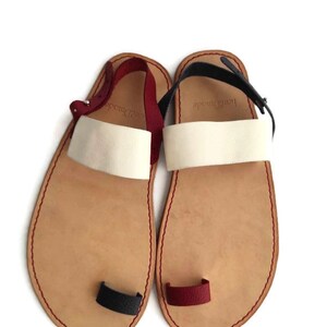 Puede incluir: Un par de sandalias de cuero con un bucle para el dedo del pie. Las sandalias son de color marr&oacute;n claro con correas blancas y detalles rojos y negros. Las sandalias tienen el texto "Hand Made" en las correas.