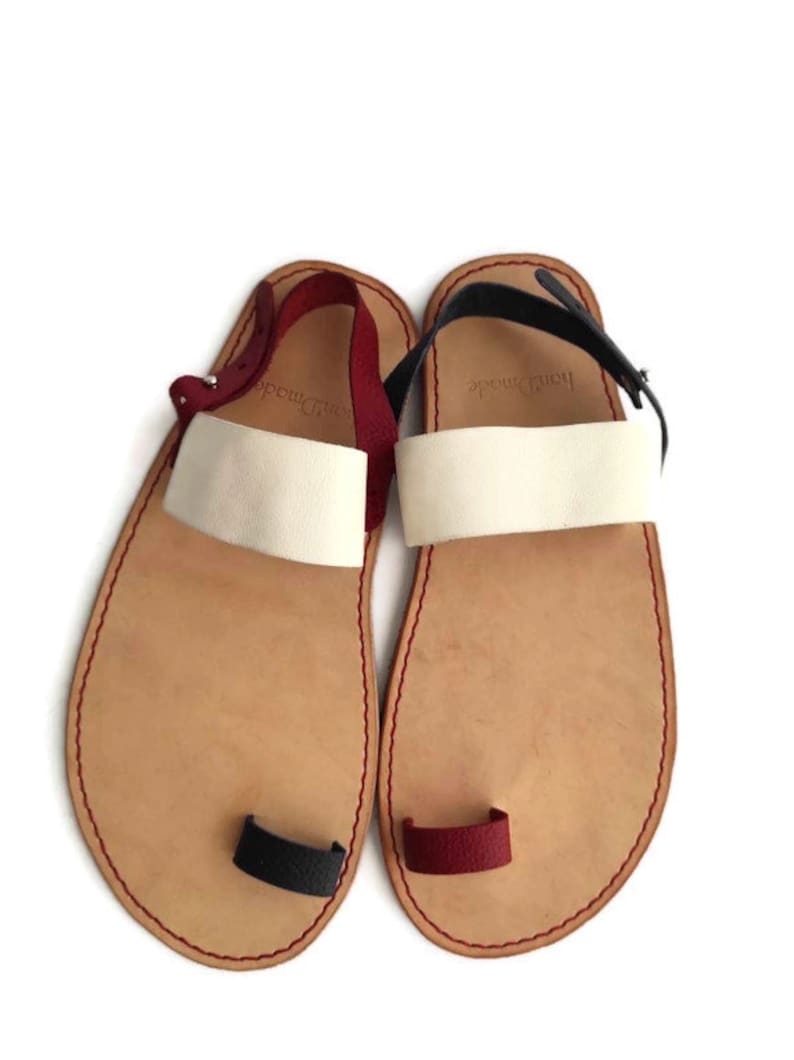 Puede incluir: Un par de sandalias de cuero con un bucle para el dedo del pie. Las sandalias son de color marr&oacute;n claro con correas blancas y detalles rojos y negros. Las sandalias tienen el texto "Hand Made" en las correas.