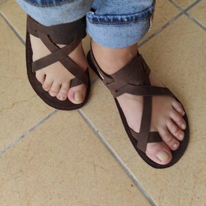 Op de afbeelding: Een paar bruine leren sandalen met gekruiste banden. De sandalen worden gedragen aan de voeten van een persoon, de tenen zijn zichtbaar.