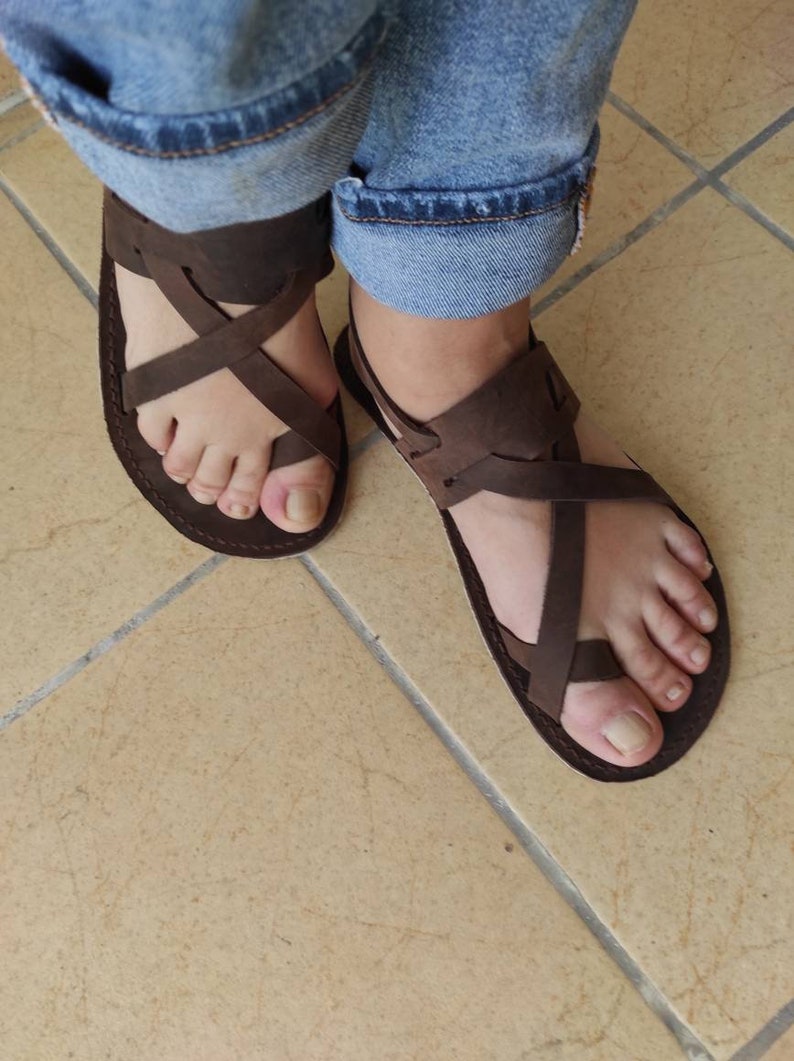 Op de afbeelding: Een paar bruine leren sandalen met gekruiste banden. De sandalen worden gedragen aan de voeten van een persoon, de tenen zijn zichtbaar.