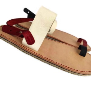 Puede incluir: Un par de sandalias de cuero con una correa blanca y una correa roja. Las sandalias tienen una suela de cuero marr&oacute;n y una puntada roja alrededor del borde.
