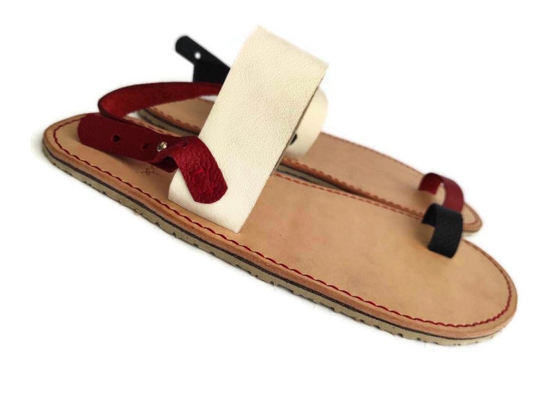 Puede incluir: Un par de sandalias de cuero con una correa blanca y una correa roja. Las sandalias tienen una suela de cuero marr&oacute;n y una puntada roja alrededor del borde.