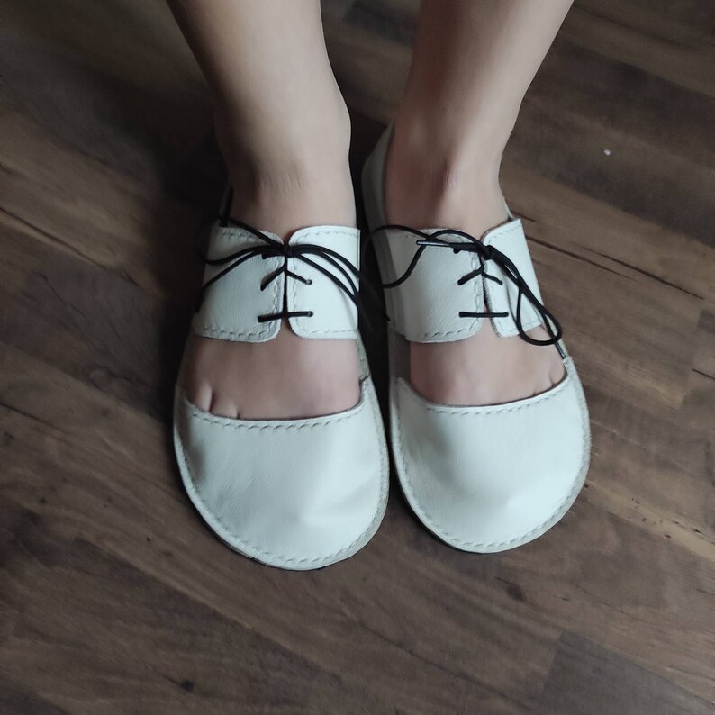 Puede incluir: Un par de sandalias de cuero blanco con cordones negros. Las sandalias tienen la punta cerrada y el tal&oacute;n abierto.