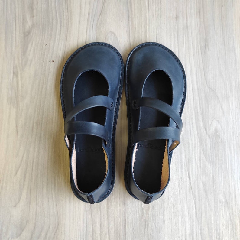 Puede incluir: Un par de zapatos planos de cuero negro con un dise&ntilde;o de doble correa. Los zapatos tienen una punta redonda y una suela plana.