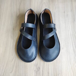 Puede incluir: Un par de zapatos planos de cuero negro con un dise&ntilde;o de correa cruzada.