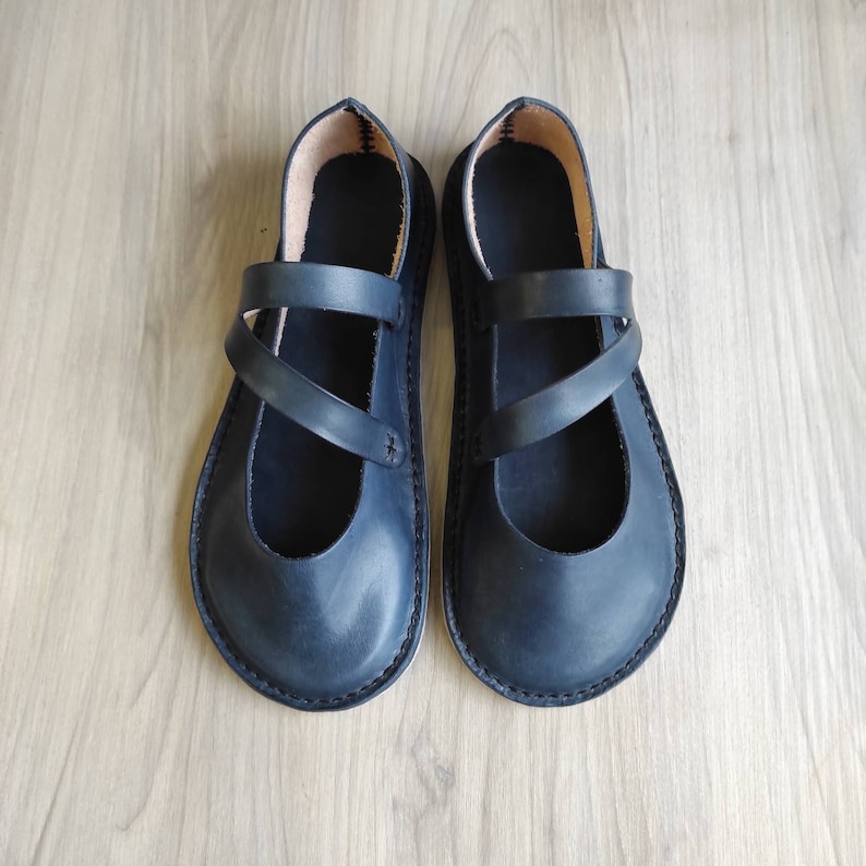 Puede incluir: Un par de zapatos planos de cuero negro con un dise&ntilde;o de correa cruzada.