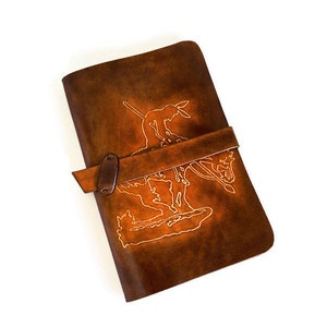 May include: A brown leather journal with a strap closure and a design of a person riding a horse on the front cover.