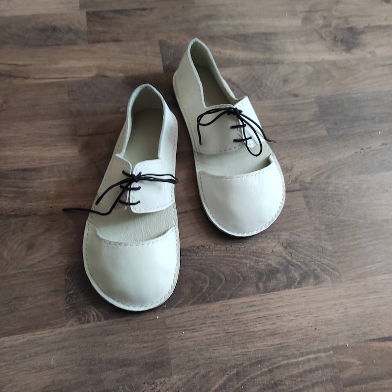 Puede incluir: Un par de sandalias de cuero blanco con cordones negros. Las sandalias tienen una punta redonda y un dorso abierto.