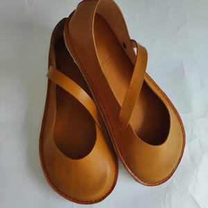 Pode incluir: Um par de sapatos baixos de couro marrom com um detalhe de costura vermelha ao redor das bordas. Os sapatos t&ecirc;m um design simples com uma &uacute;nica tira na parte superior.