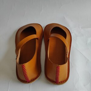 Pode incluir: Um par de sapatos de couro castanho com costuras vermelhas ao redor das bordas. Os sapatos t&ecirc;m uma tira na parte superior e uma ponta arredondada.