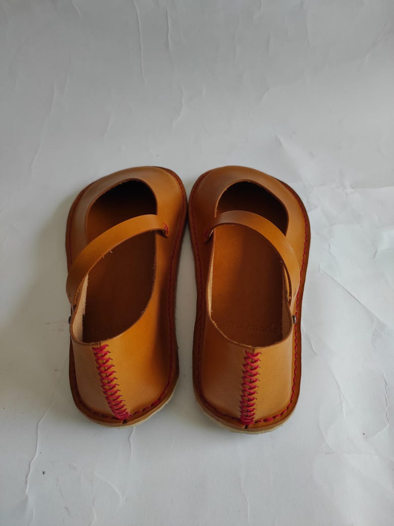 Pode incluir: Um par de sapatos de couro castanho com costuras vermelhas ao redor das bordas. Os sapatos t&ecirc;m uma tira na parte superior e uma ponta arredondada.