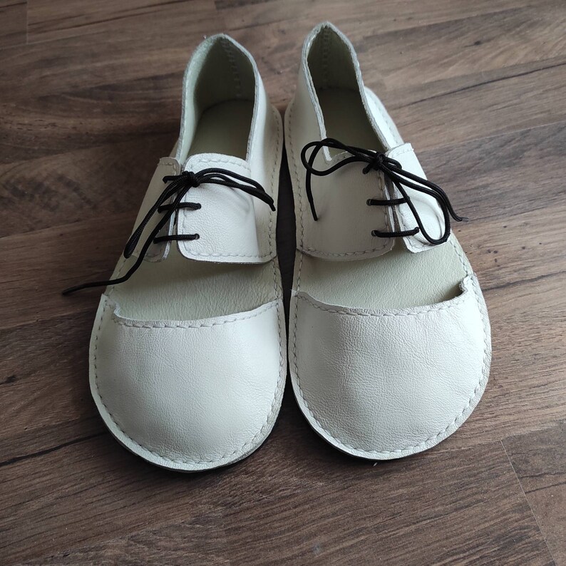 Puede incluir: Un par de zapatos blancos de cuero con cordones negros. Los zapatos tienen una punta redonda y un dise&ntilde;o sencillo.