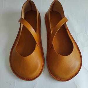 Pode incluir: Um par de sapatos baixos de couro marrom com uma &uacute;nica tira na parte superior. Os sapatos t&ecirc;m costuras vermelhas ao redor das bordas.