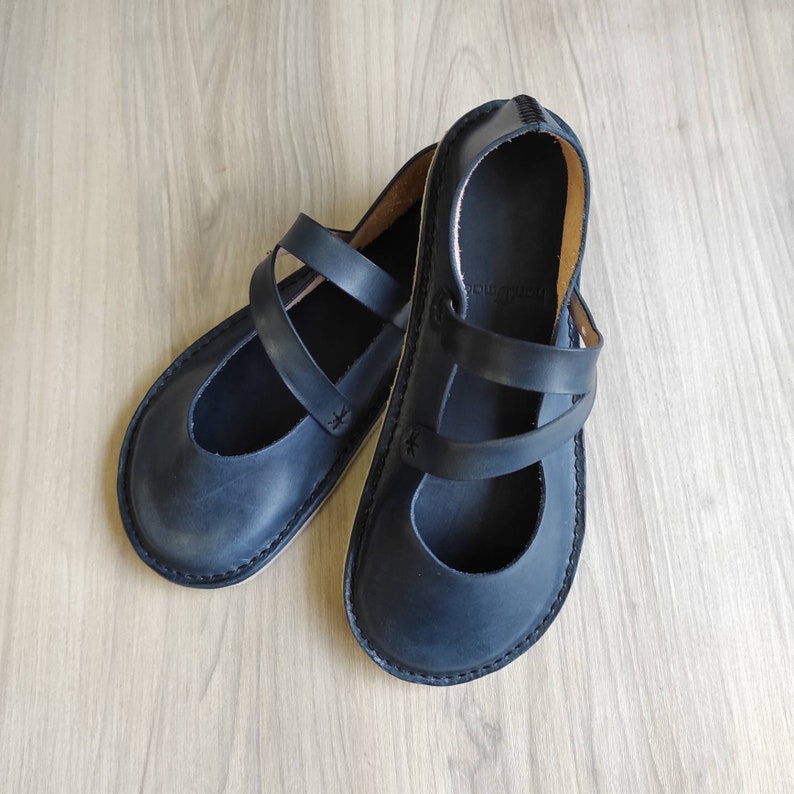 Puede incluir: Un par de sandalias de cuero negro con un dise&ntilde;o de correa. Las sandalias tienen una punta redonda y una suela plana.