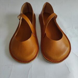 Pode incluir: Um par de sapatos baixos de couro marrom com costuras vermelhas ao redor das bordas. Os sapatos t&ecirc;m um design simples com uma &uacute;nica tira na parte superior.
