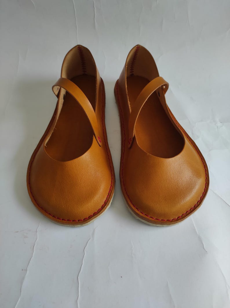 Pode incluir: Um par de sapatos baixos de couro marrom com costuras vermelhas ao redor das bordas. Os sapatos t&ecirc;m um design simples com uma &uacute;nica tira na parte superior.