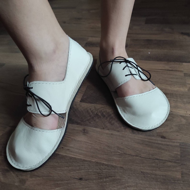 Puede incluir: Sandalias de cuero blanco con cordones negros. Las sandalias tienen un dise&ntilde;o recortado y una punta redonda.