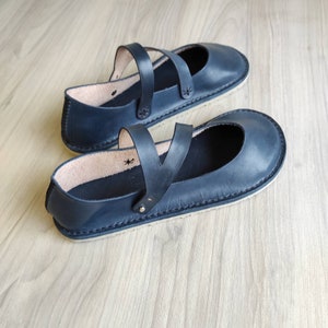 Puede incluir: Un par de zapatos planos de cuero azul oscuro con una correa en la parte superior. Los zapatos tienen un dise&ntilde;o simple y un aspecto c&oacute;modo.