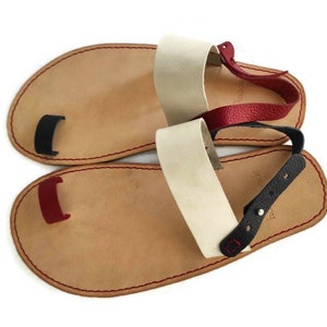 Puede incluir: Un par de sandalias de cuero con un bucle para los dedos. Las sandalias tienen una base de cuero marr&oacute;n con correas de cuero rojo, negro y blanco.