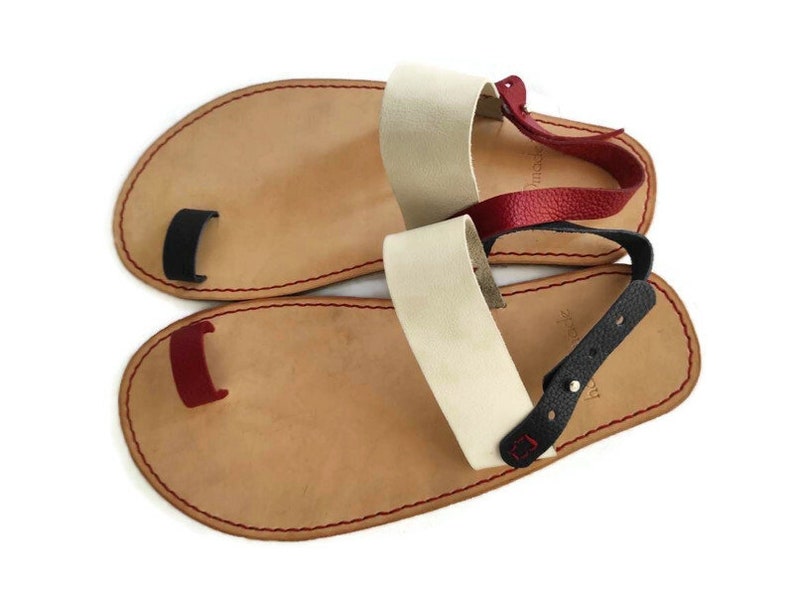 Puede incluir: Un par de sandalias de cuero con un bucle para los dedos. Las sandalias tienen una base de cuero marr&oacute;n con correas de cuero rojo, negro y blanco.