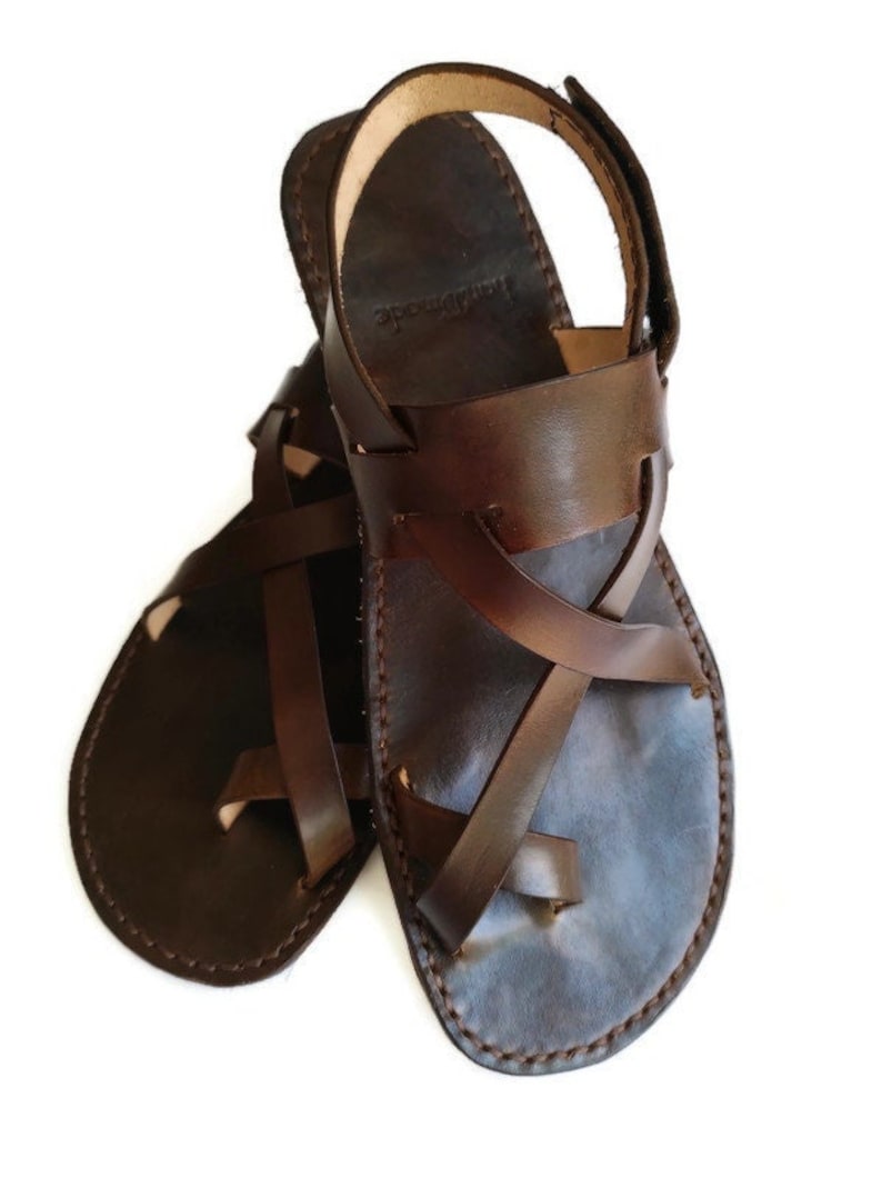 Op de afbeelding: Een paar bruine leren sandalen met gekruiste banden en een handgemaakt label. De sandalen hebben een gesloten teen en een open hiel.