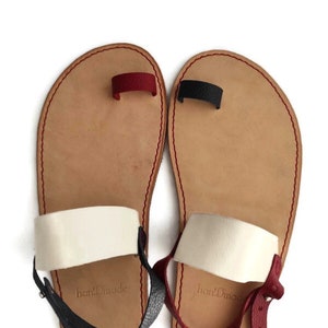 Puede incluir: Un par de sandalias de cuero marr&oacute;n con correas blancas y rojas. Las sandalias tienen un bucle para el dedo del pie y una correa para el tobillo con una hebilla. Las sandalias est&aacute;n hechas a mano y tienen el texto "handmade" en las correas.