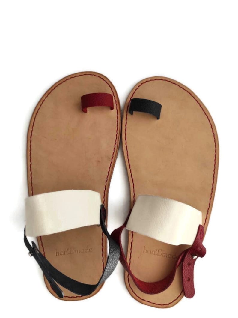 Puede incluir: Un par de sandalias de cuero marr&oacute;n con correas blancas y rojas. Las sandalias tienen un bucle para el dedo del pie y una correa para el tobillo con una hebilla. Las sandalias est&aacute;n hechas a mano y tienen el texto "handmade" en las correas.