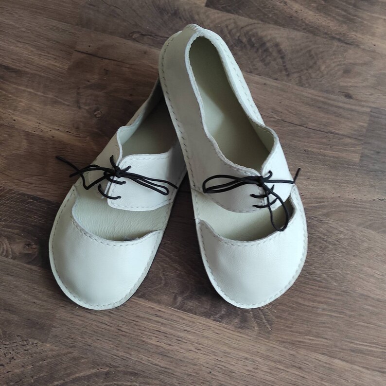 Puede incluir: Un par de zapatos de cuero blanco con cordones negros. Los zapatos tienen un dise&ntilde;o recortado en la parte superior y una punta redonda.