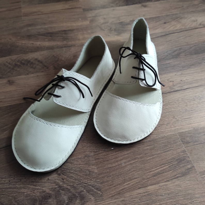 Puede incluir: Un par de zapatos de cuero blanco con cordones negros. Los zapatos tienen un dise&ntilde;o recortado en la parte superior y una punta redonda.