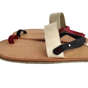 Puede incluir: Un par de sandalias de cuero con una trabilla para el dedo gordo y una correa para el tobillo. Las sandalias est&aacute;n hechas de cuero marr&oacute;n con costuras rojas. La correa del tobillo est&aacute; hecha de cuero blanco con una correa de cuero negro y una estrella roja.