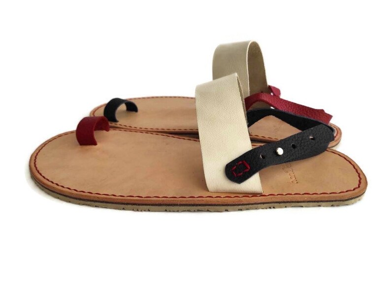 Puede incluir: Un par de sandalias de cuero con una trabilla para el dedo gordo y una correa para el tobillo. Las sandalias est&aacute;n hechas de cuero marr&oacute;n con costuras rojas. La correa del tobillo est&aacute; hecha de cuero blanco con una correa de cuero negro y una estrella roja.