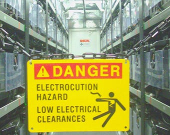 May include: A yellow danger sign with red text reading "DANGER" and the words "ELECTROCUTION HAZARD" and "LOW ELECTRICAL CLEARANCES". The sign features a stick figure being electrocuted. The sign is hanging in a room with rows of equipment.