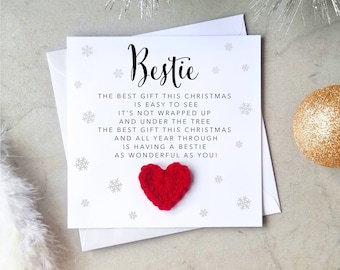 Könnte beinhalten: Weiße Grußkarte mit dem Wort "Bestie" in Schreibschrift. Die Karte zeigt ein rotes Herz und Schneeflocken, mit einer Botschaft über Freundschaft. Eine goldene Kugel und silberne Zierzweige sind im Hintergrund.