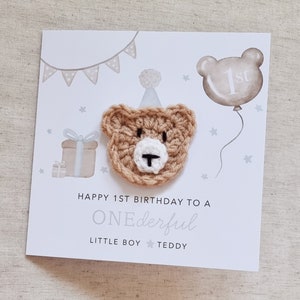 Puede incluir: Una tarjeta blanca con un oso de crochet marr&oacute;n que lleva un gorro de fiesta blanco. La tarjeta dice "Happy 1st Birthday to a ONEderful Little Boy Teddy".