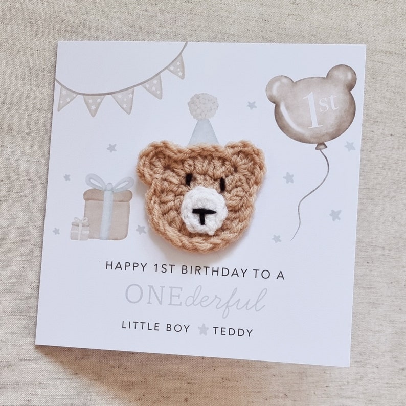Puede incluir: Una tarjeta blanca con un oso de crochet marr&oacute;n que lleva un gorro de fiesta blanco. La tarjeta dice "Happy 1st Birthday to a ONEderful Little Boy Teddy".