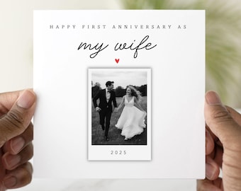 May include: A white card with the text "HAPPY FIRST ANNIVERSARY AS my wife" in black script. A small red heart is below the text. A black and white photo of a couple is in the center, with the year 2025 below the photo.