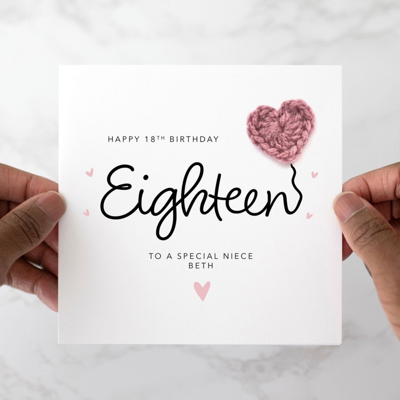 Puede incluir: Una tarjeta blanca con el texto "Happy 18th Birthday" y "Eighteen" en letra cursiva negra. La tarjeta tambi&eacute;n tiene el texto "To a special niece Beth" y un coraz&oacute;n rosa de ganchillo.