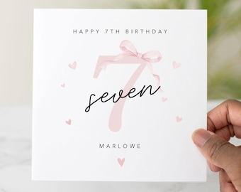 May include: A white birthday card with the text "HAPPY 7TH BIRTHDAY" at the top. A large pink number 7 is in the center, with the word "seven" written below it in cursive. Small pink hearts are scattered around the card.