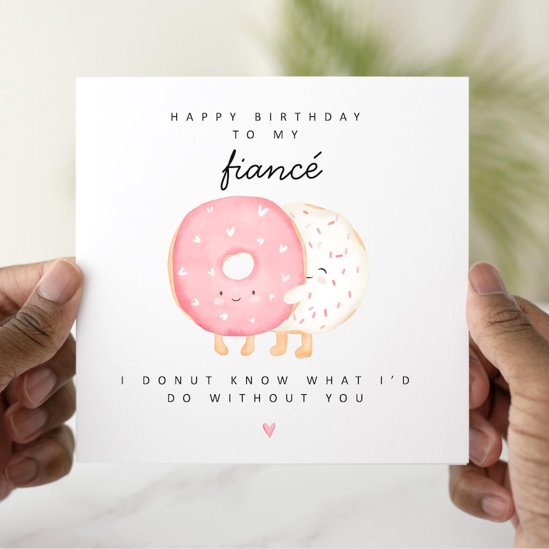 Op de afbeelding: Een witte kaart met een roze en witte donut-illustratie. De kaart luidt "Happy Birthday aan mijn verloofde. Ik weet niet wat ik zonder jou zou doen."