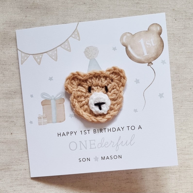 Puede incluir: Una tarjeta blanca con un oso de crochet marr&oacute;n que lleva un gorro de fiesta. La tarjeta dice "Happy 1st Birthday to a ONEderful SON MASON".