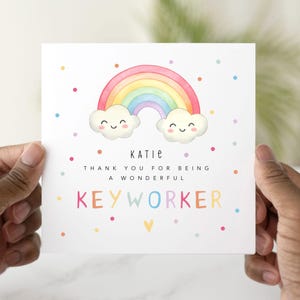 May include: A white card with a rainbow and two clouds with smiley faces. The text on the card reads "Katie, Thank you for being a wonderful Keyworker."