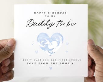 May include: A white greeting card held by hands, with the text "HAPPY BIRTHDAY TO MY Daddy to be". A blue watercolor heart and small blue hearts are featured. The card also reads "I CAN'T WAIT FOR OUR FIRST CUDDLE LOVE FROM THE BUMP X".