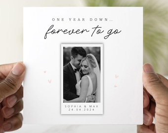 May include: A white card with the text "ONE YEAR DOWN... forever to go" in black script. A black and white photo of a couple is centered on the card, with the names "SOPHIA & MAX" and the date "24.04.2024" below the photo. Small pink hearts are scattered around the photo.
