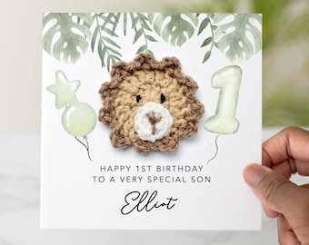 May include: A white card with green leaves printed on it. The card has a crocheted lion, a green balloon with a star, and a green number one balloon. The card says "Happy 1st Birthday to a very special son Elliot".