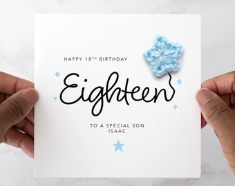 May include: White birthday card with the text "Happy 18th Birthday" and "Eighteen" in a handwritten font. A light blue crocheted star is attached to the card, along with small blue star accents. The card is held by two hands.