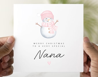 May include: A white Christmas card featuring a watercolor snowman wearing a pink hat and scarf. The card reads "MERRY CHRISTMAS TO A VERY SPECIAL Nana" in black script, with a small pink heart below the name. The card is held by hands.